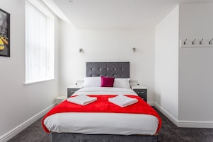 A modern hotel room with a large bed, a red throw blanket, and a grey headboard. There are two bedside tables with lamps and a window with blinds.
