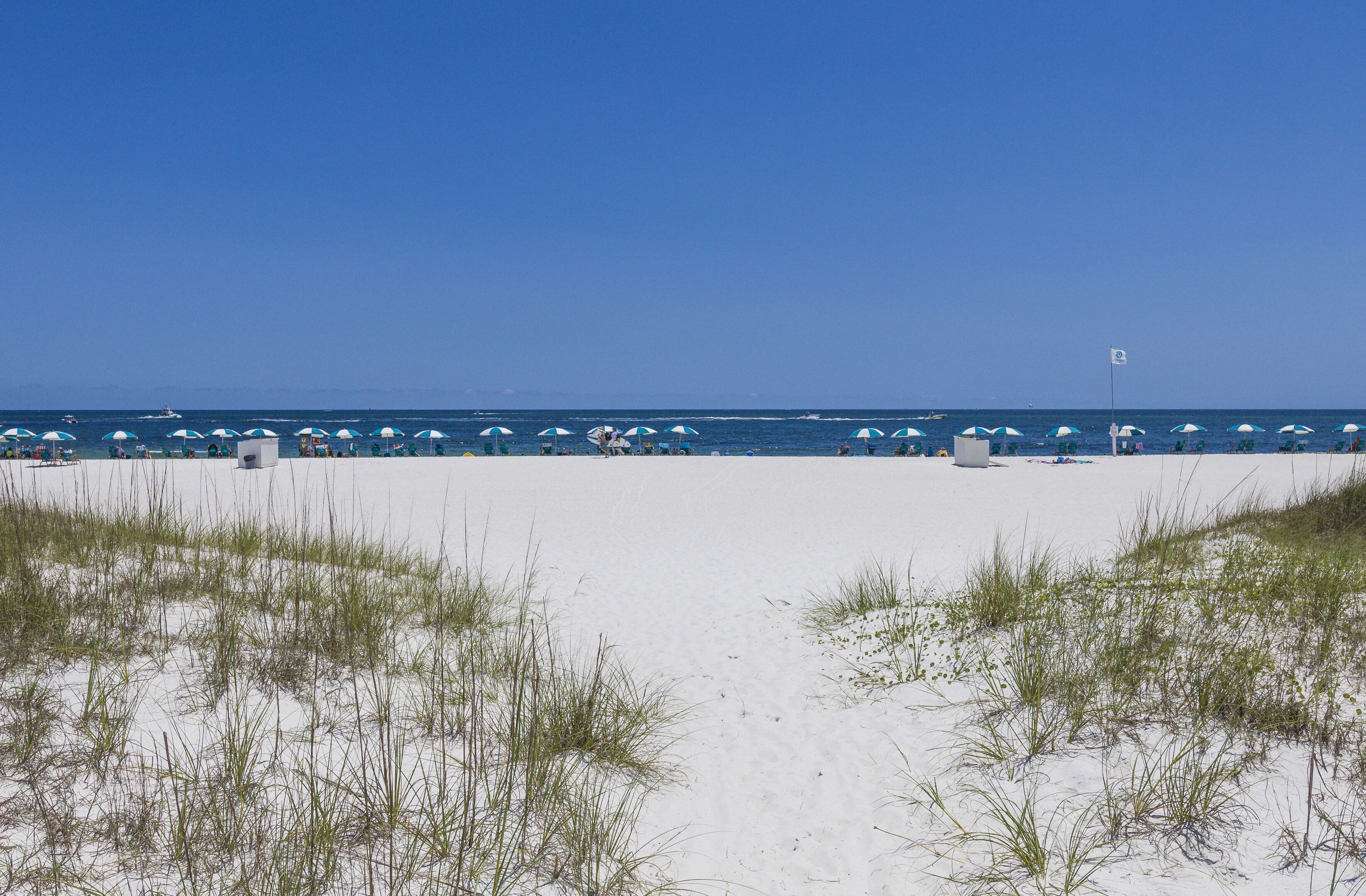Ein Strand mit weißem Sand und vereinzelten Gräsern im Vordergrund, ein klarer, blauer Himmel und eine ruhige See im Hintergrund.