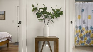 Une pièce avec un lit, un vase blanc contenant des feuilles vertes, posé sur une table d’appoint en bois, et un rideau de douche à motifs.