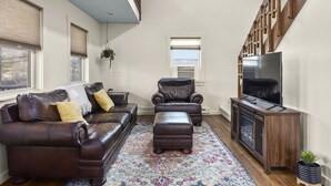 Un salon avec un canapé en cuir marron, un pouf assorti, un meuble de rangement en bois avec un téléviseur et un tapis à motifs.
