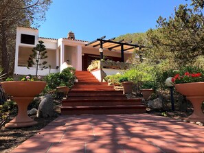 A building with a stone facade, a wooden pergola, and a paved pathway leading to a set of stairs. - Villa La Maddalena in La Maddalena (La Maddalena)