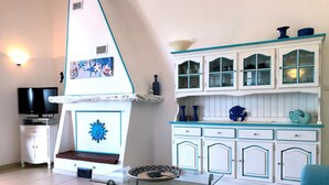 A living room with a TV, a fireplace, a dining table, and a kitchen area with cabinets. - Villa La Maddalena in La Maddalena (La Maddalena)