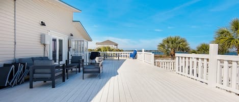 A wooden deck with outdoor seating, a white building, and a view of the ocean.