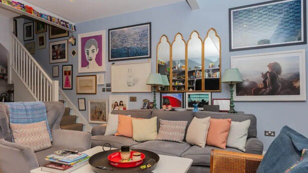 A living room with a grey sofa, a coffee table with books and a tray, a staircase with framed pictures, and a bookshelf with various items.