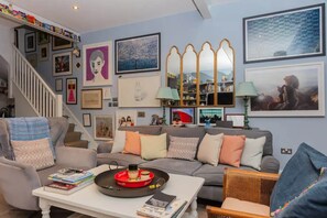 Una sala de estar con un sofá gris, una mesita de centro con libros y una bandeja, una escalera con cuadros enmarcados y una estantería con diversos objetos.