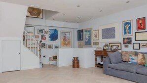 A spacious living room with a grey sofa, a wooden coffee table, and a staircase leading to another level. The walls are adorned with various framed artworks and a large clock.