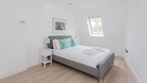 A modern bedroom with a grey bed, white walls, a small window, and a nightstand.