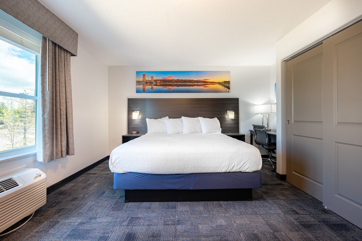 A hotel room with a large bed, a desk, a chair, a window with curtains, and a painting of a bridge.