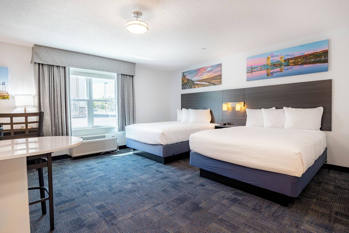 A hotel room with two beds, a desk, a chair, a window with curtains, and two wall-mounted paintings.