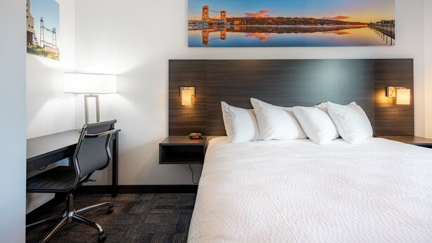 A hotel room with a large bed, a desk with a chair, a nightstand with a lamp, and a painting of a bridge on the wall. - Independence Stay (Houghton)