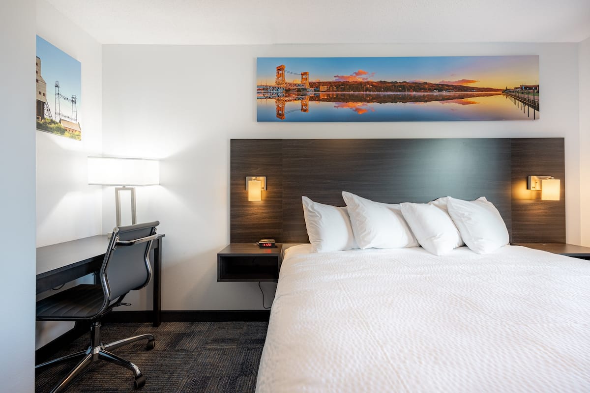 A hotel room with a large bed, a desk with a chair, a nightstand with a lamp, and a painting of a bridge on the wall.