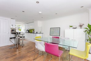 A modern kitchen with white cabinets, a glass dining table, and colorful chairs. - Stylish 4BDR Home 3 mins from Kings Cross w Garden (London)