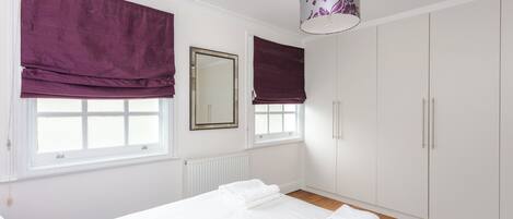 Un dormitorio con cama, dos ventanas con cortinas moradas, un espejo, un armario y una lámpara colgante.
