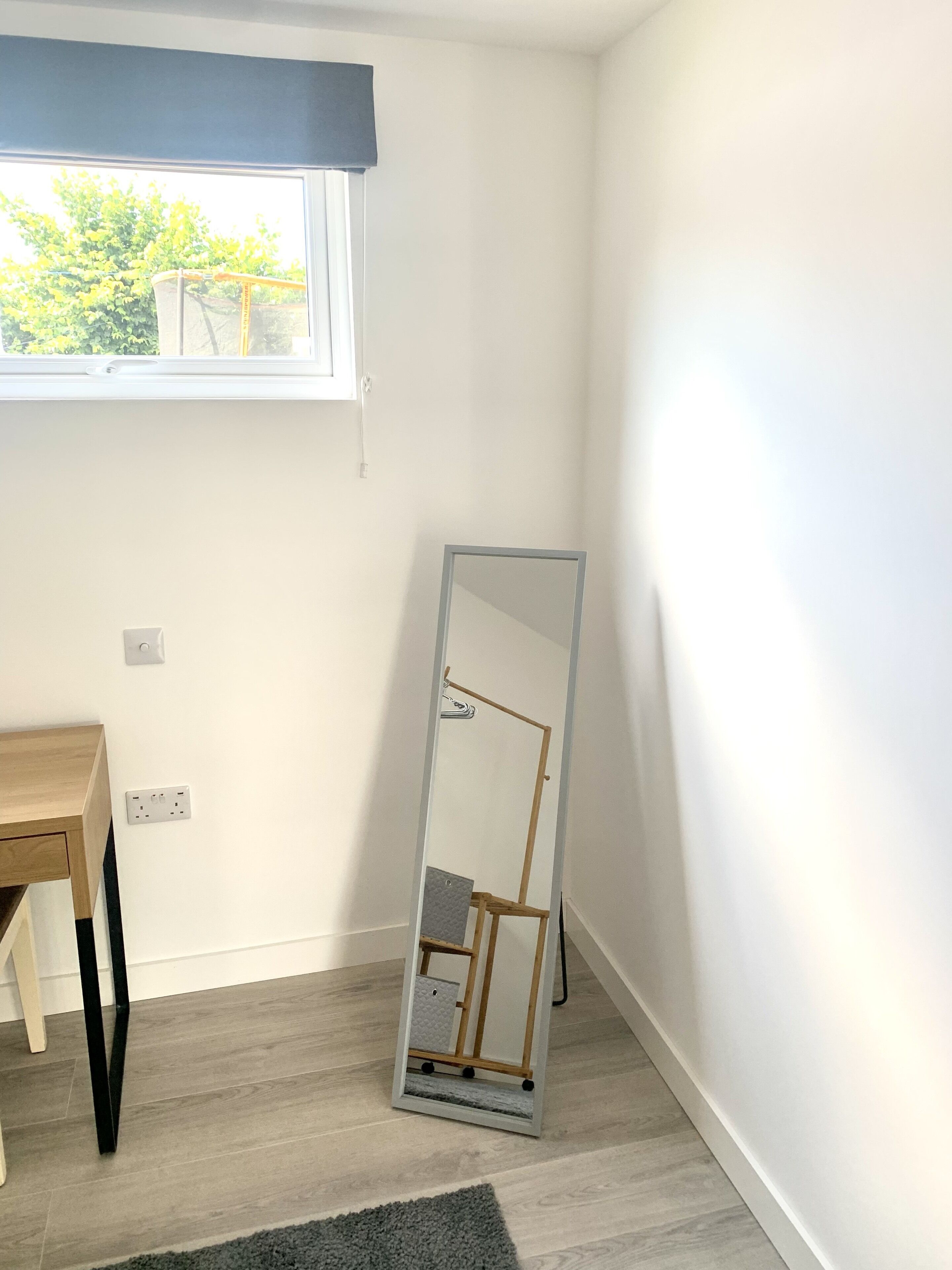 A room with a window, a mirror, a wooden table, and a grey rug.