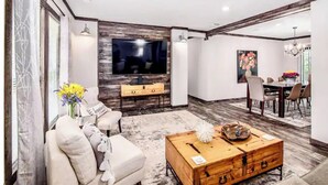 Una sala moderna con una televisión de pantalla plana montada en una pared texturizada, una mesa de centro de madera, un sofá y una zona de comedor con una araña.