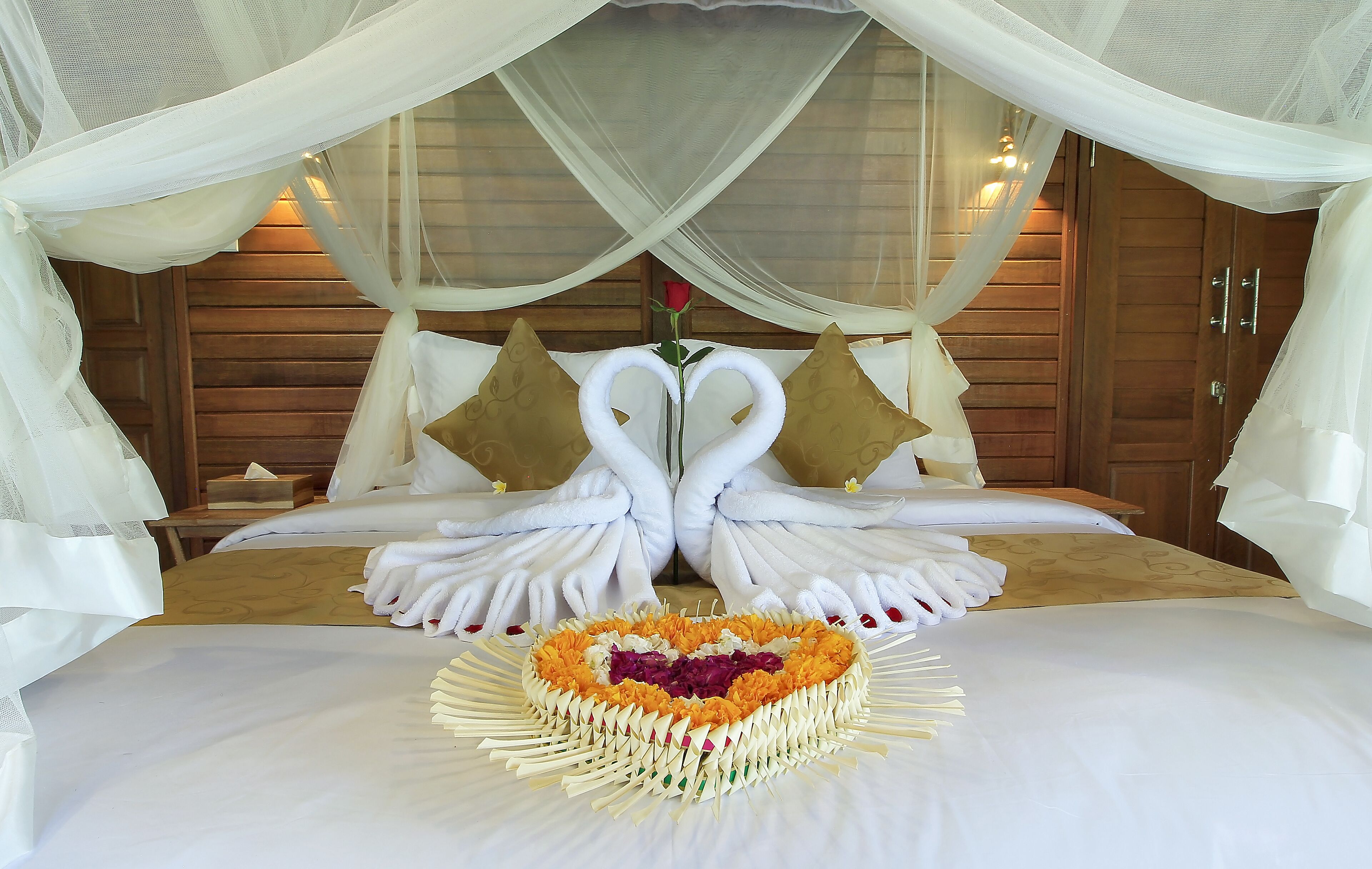 Une chambre d’hôtel luxueuse avec un lit à baldaquin, un arrangement floral en forme de cœur et des serviettes soigneusement pliées.