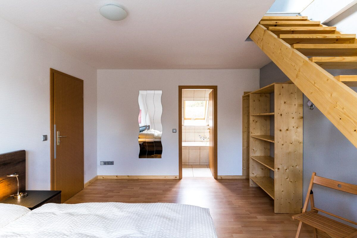 A bedroom with a wooden bed, a nightstand, a wooden chair, a wooden staircase, and a view of a bathroom through a doorway.