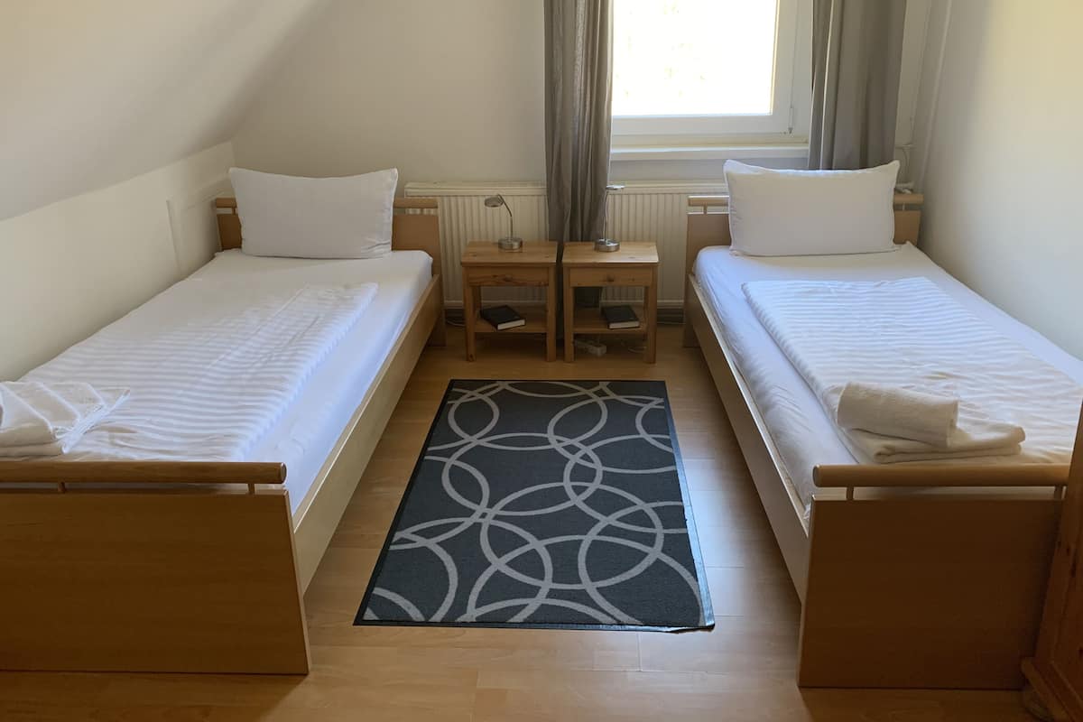Une chambre double avec deux lits simples, une petite table en bois avec une lampe, un radiateur et une fenêtre avec des rideaux.