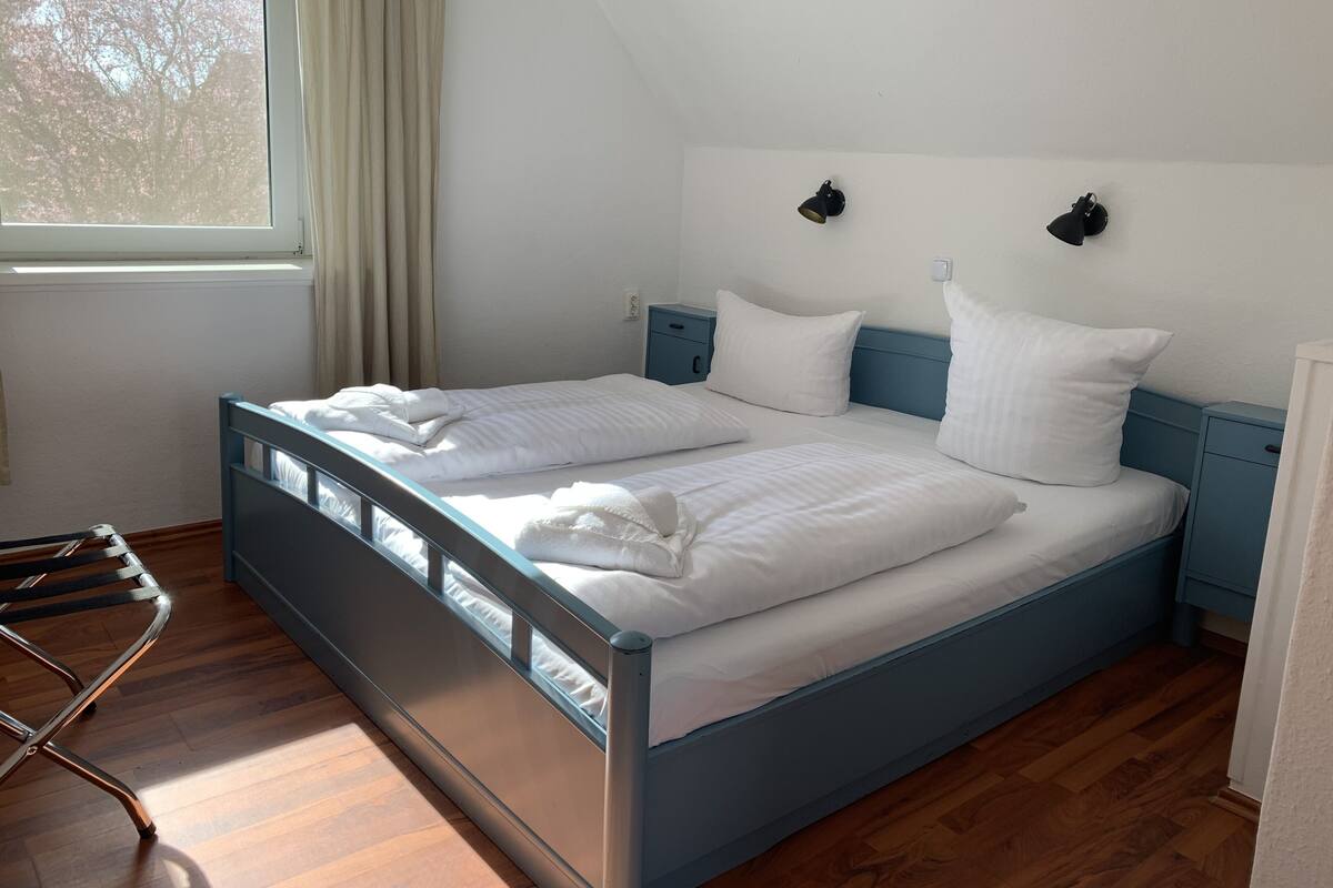 Un lit double avec du linge de lit blanc, un plancher en bois, une tête de lit bleue et une table de chevet avec une lampe.