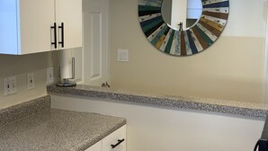 A kitchen with white cabinets, a granite countertop, a round mirror with a multicolored frame, and a door leading to another room.