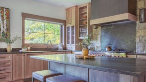 A modern kitchen with wooden cabinets, a large island with stools, a stone accent wall, and a window with a view of trees. - Dreamy home on river, mountain views, steps from town. (Frisco)