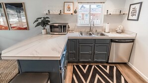 Een compacte keuken met een marmeren aanrecht, donkere kastjes, een magnetron, een spoelbak en een vaatwasser. Boven het aanrecht bevinden zich planken met glazen en een potplant.