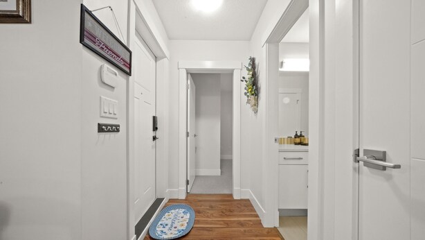 A hallway with white walls, wooden flooring, and a welcome mat. There are light switches and a framed picture on the wall.