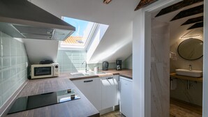 Ett kompakt kök med mikrovågsugn, kaffebryggare och diskmaskin. Ett takfönster ger naturligt ljus, och köket har anslutning till ett badrum med handfat och spegel.