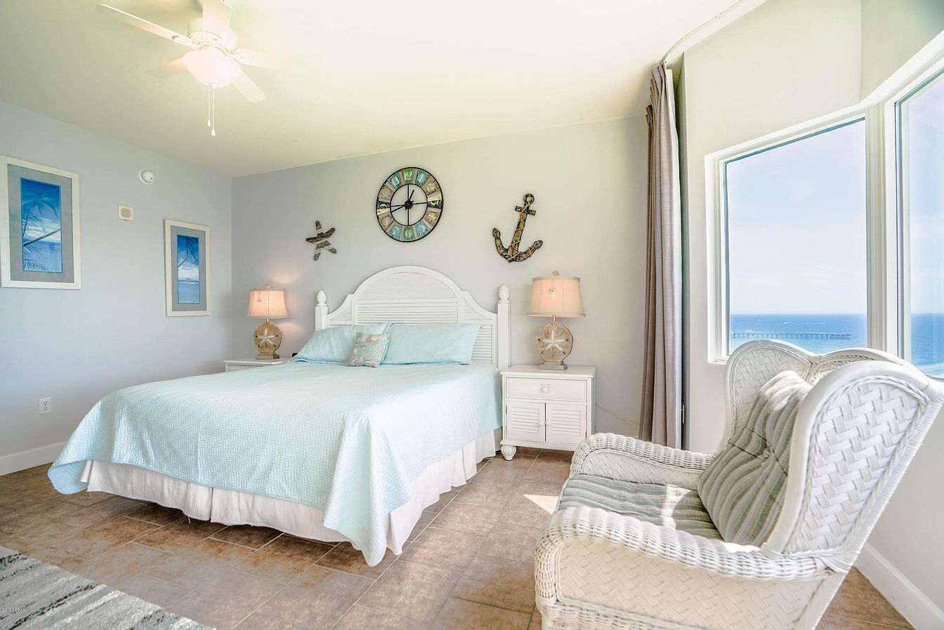 A bedroom with a bed, a nightstand, a chair, a ceiling fan, a wall clock, and a window with a view of the ocean.