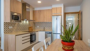 A modern kitchen with wooden cabinets, a microwave, a stove, and a refrigerator. There is a dining table with chairs and a potted plant. - HomeHolidaysRentals Benoit - Costa Barcelona (Pineda de Mar)