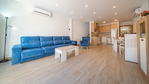 A modern living room with a blue sofa, a white coffee table, and a blue armchair. The kitchen is visible in the background with wooden cabinets and a white refrigerator. - HomeHolidaysRentals Benoit - Costa Barcelona (Pineda de Mar)