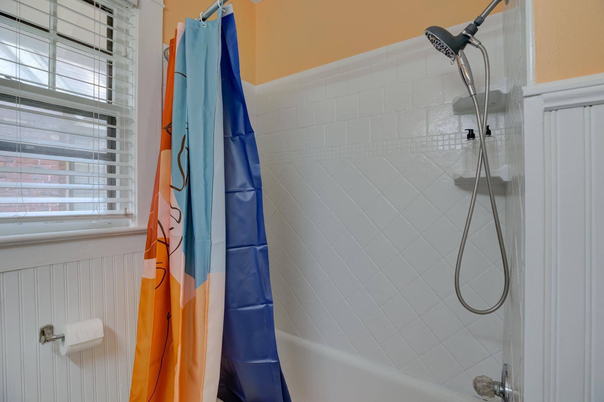 A bathroom with a shower, white tiled walls, a window with blinds, and a colorful shower curtain.