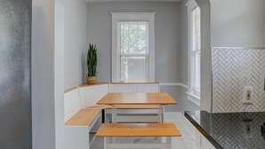 Una cocina con mesa y banco de madera, armarios blancos, un ventanal con persianas y salpicadero con motivo de espiga.