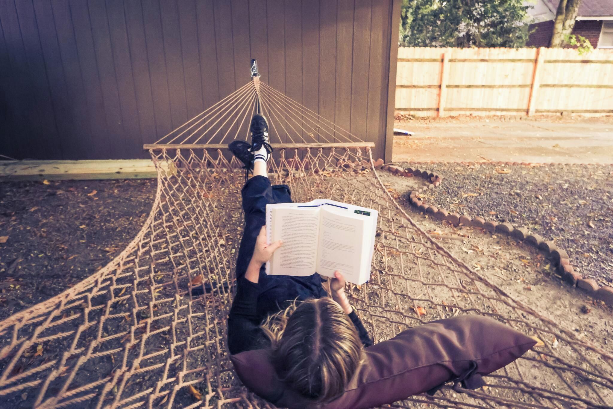 A hammock with a person reading a book, set against a wooden fence and a building.