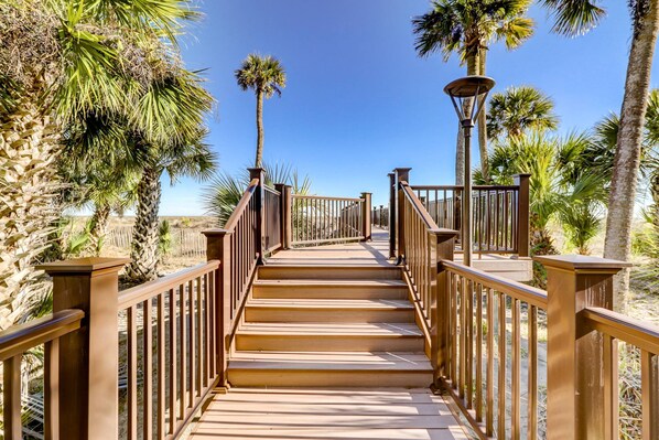 A wooden boardwalk leading to a beach, surrounded by palm trees and a clear blue sky.