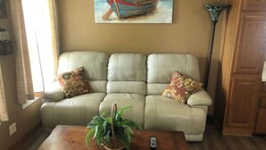 Un salon avec un canapé beige, une table basse en bois ornée d’une plante, une télécommande et un tableau représentant un bateau accroché au mur.