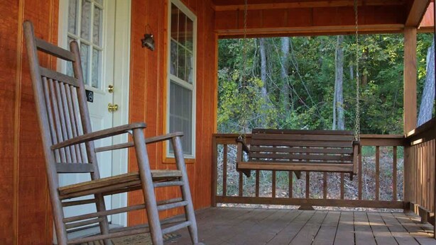 Une véranda en bois avec un fauteuil à bascule et un banc, une porte et une vue sur les arbres.