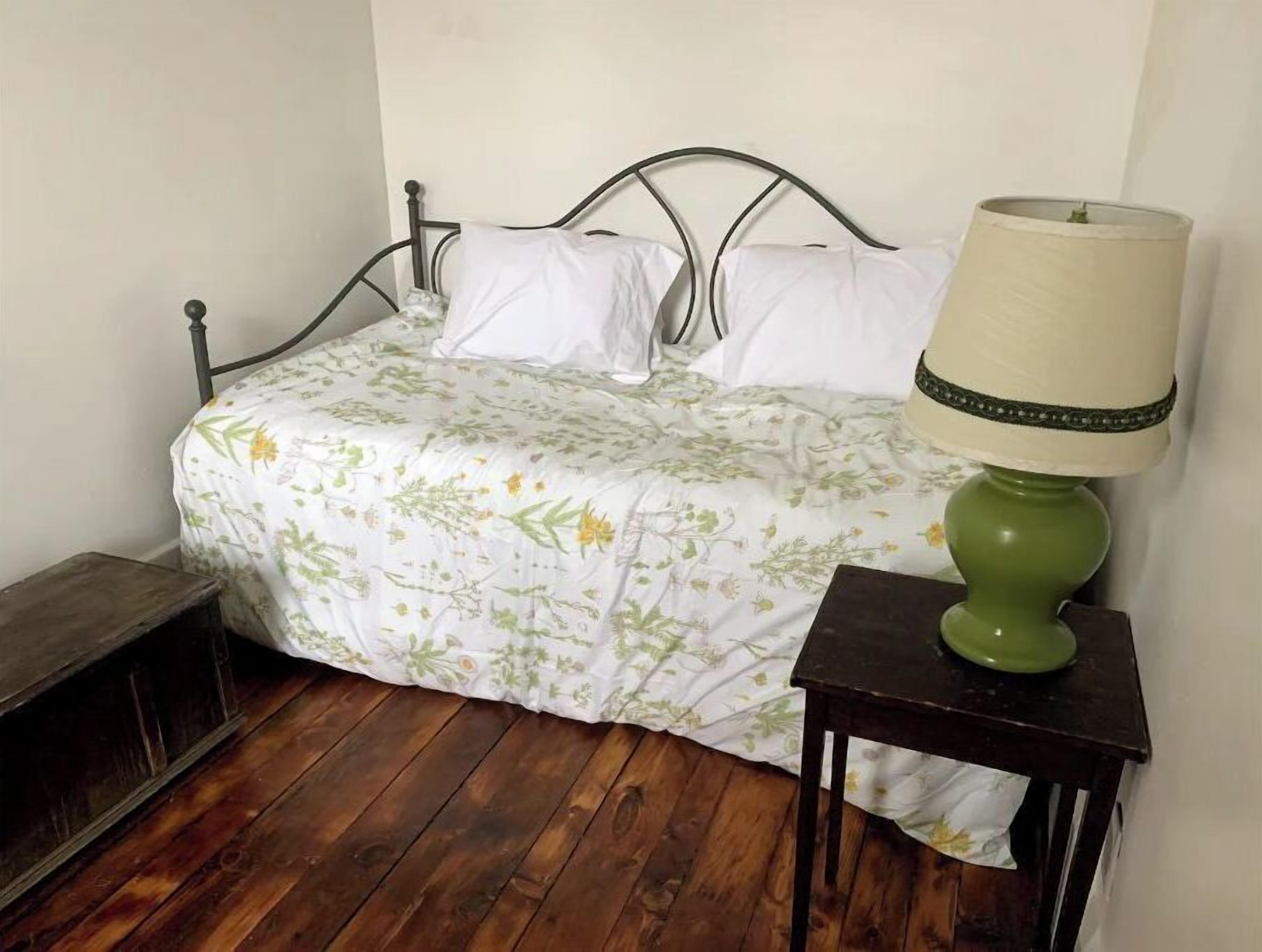 Un lit recouvert d’une couvre-lit à motifs floraux, une lampe verte posée sur une table de chevet et un banc en bois.