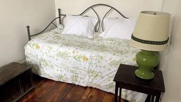 Un lit recouvert d’une couvre-lit à motifs floraux, une lampe verte posée sur une table de chevet et un banc en bois.