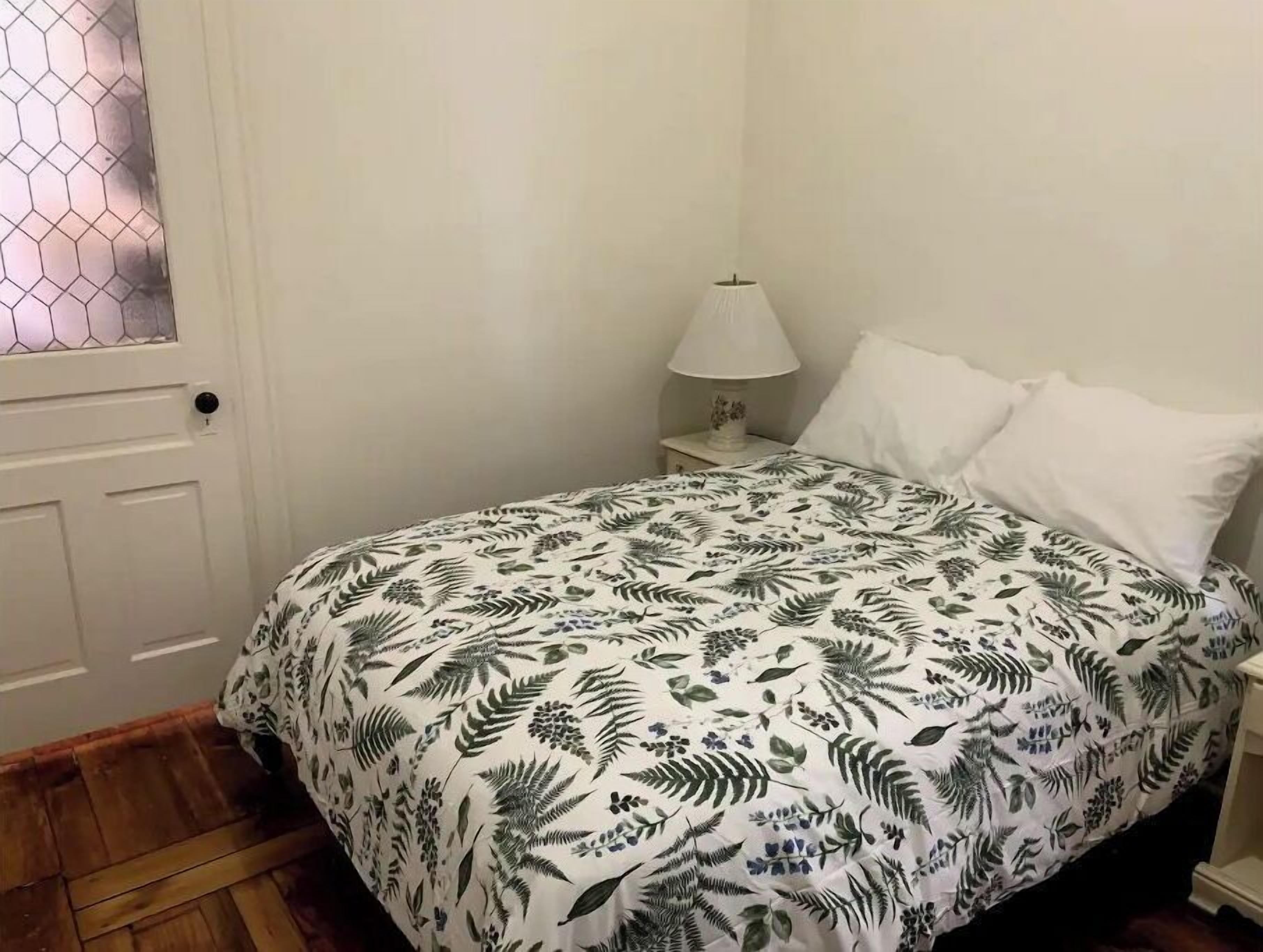 Un dormitorio con cama, ropa de cama blanca, una mesita de noche con lámpara y una puerta con ventana de vidrio esmerilado.
