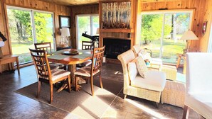 A wooden interior with a dining table, chairs, a fireplace, and large windows.