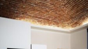 Una stanza con soffitto in mattoni, pareti bianche, una TV a schermo piatto fissata al muro, una scrivania bianca e pavimento in legno.