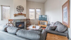 Een woonkamer met een stenen open haard, grijze banken, een salontafel en een televisie op een houten dressoir.