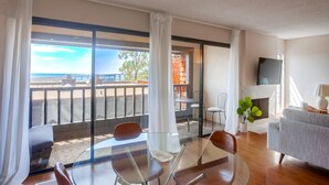 Un salon moderne avec une table en verre, des chaises, un canapé gris, un téléviseur et un balcon offrant une vue sur la plage.