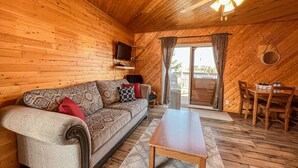 Et hyggeligt sommerhusinteriør med træloft, sofa med mønstrede puder, et træbord og et spiseområde med stole.