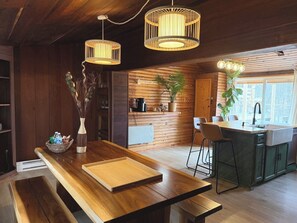 A wooden interior with a wooden table, bench, hanging light fixtures, and a kitchen area with a sink and stools.