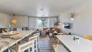Ein modernes Wohnzimmer mit Essbereich, einer Sofagarnitur, einem an der Wand befestigten Fernseher und einem großen Fenster mit Vorhängen.