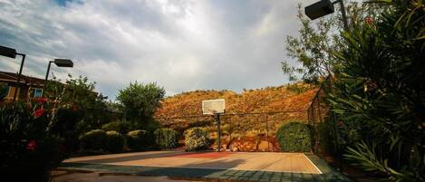Una cancha de baloncesto al aire libre con un aro, rodeada de vegetación y un paisaje montañoso al fondo.