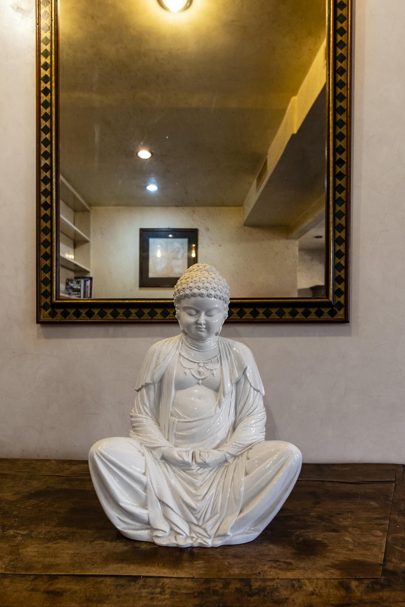 Una estàtua blanca d'una figura asseguda en posició meditativa, col·locada sobre un terra de fusta amb un gran mirall a sobre.