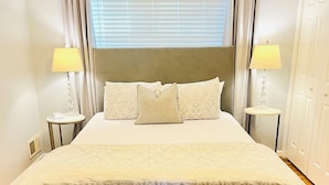 Una cama grande con cabecera, dos mesitas de noche con lámparas, una ventana con persianas y suelo de madera.